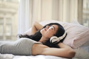 Woman laid on a bed, headphones on, eyes closed, smiling.